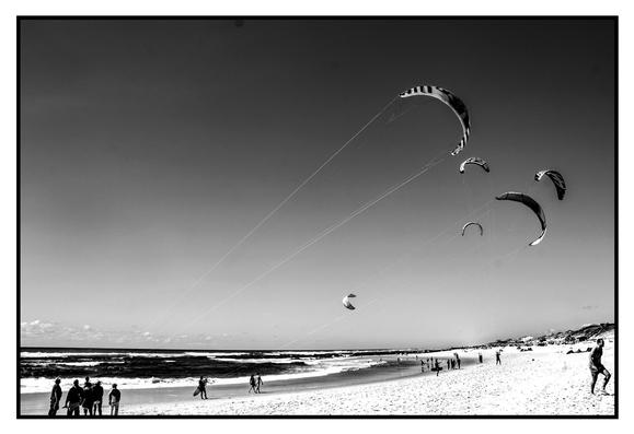 Schwarzweissbild mit hohem Kontrast. Man sieht einen weiten Sandstrand mit Badegästen und vielen Kitesurfen. Einer zerschneidet den Himmel diagonal von links unten nach rechts oben.