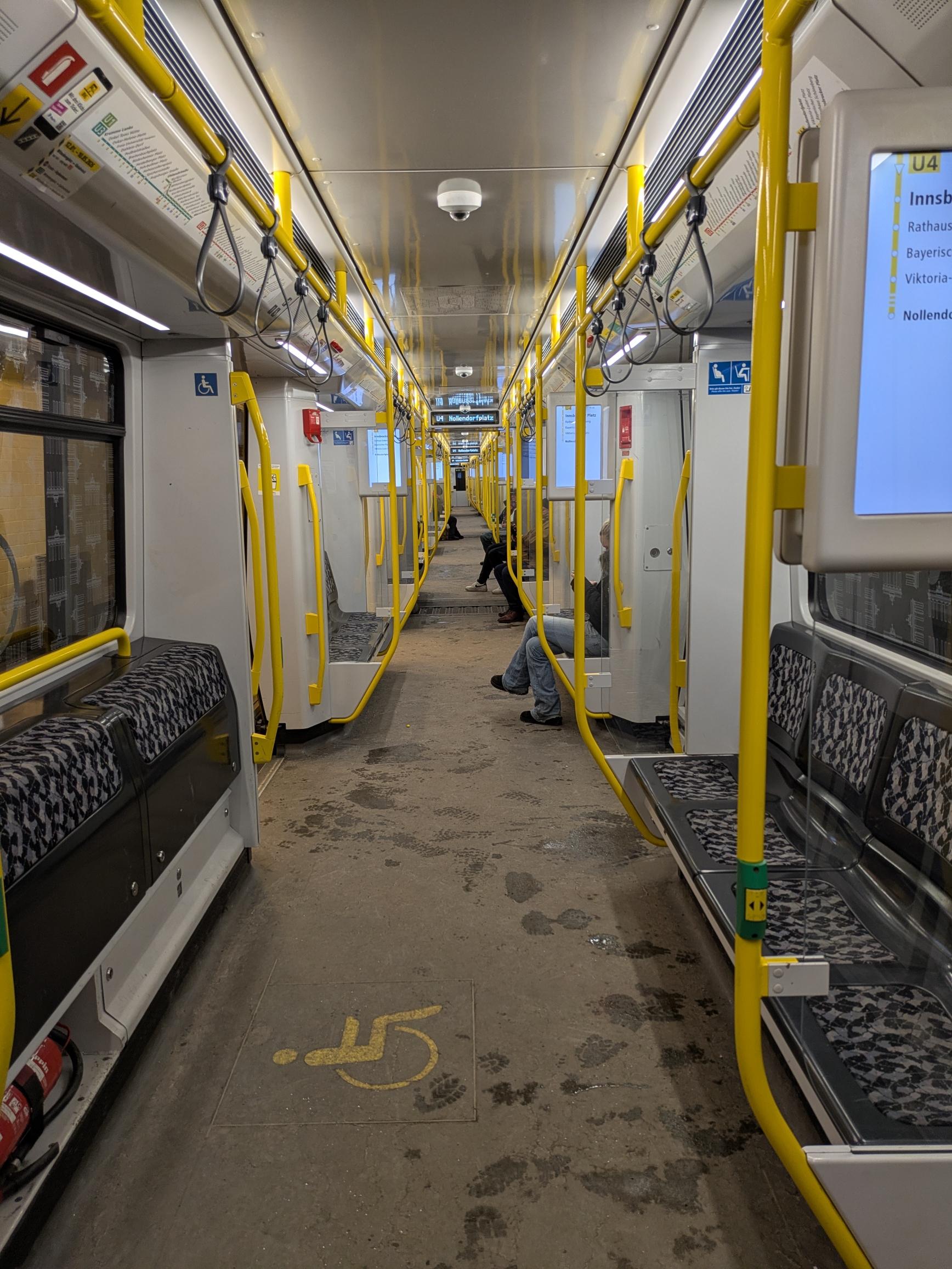 Blick längs durch einen U-Bahn Zug, man sieht somit bis zum Ende durchs, links wie rechts sind Sitze mit gelben Haltestangen. Die Wände und Decken sind weiß-grau ausgestaltet. Man sieht ebenfalls ein paar Monitore mit den nächsten Stationen. Der Boden ist grau und mit einigen Fußspuren von Wasser. Außerdem sitzen einige Personen in der Bahn, dessen Beine sieht man.