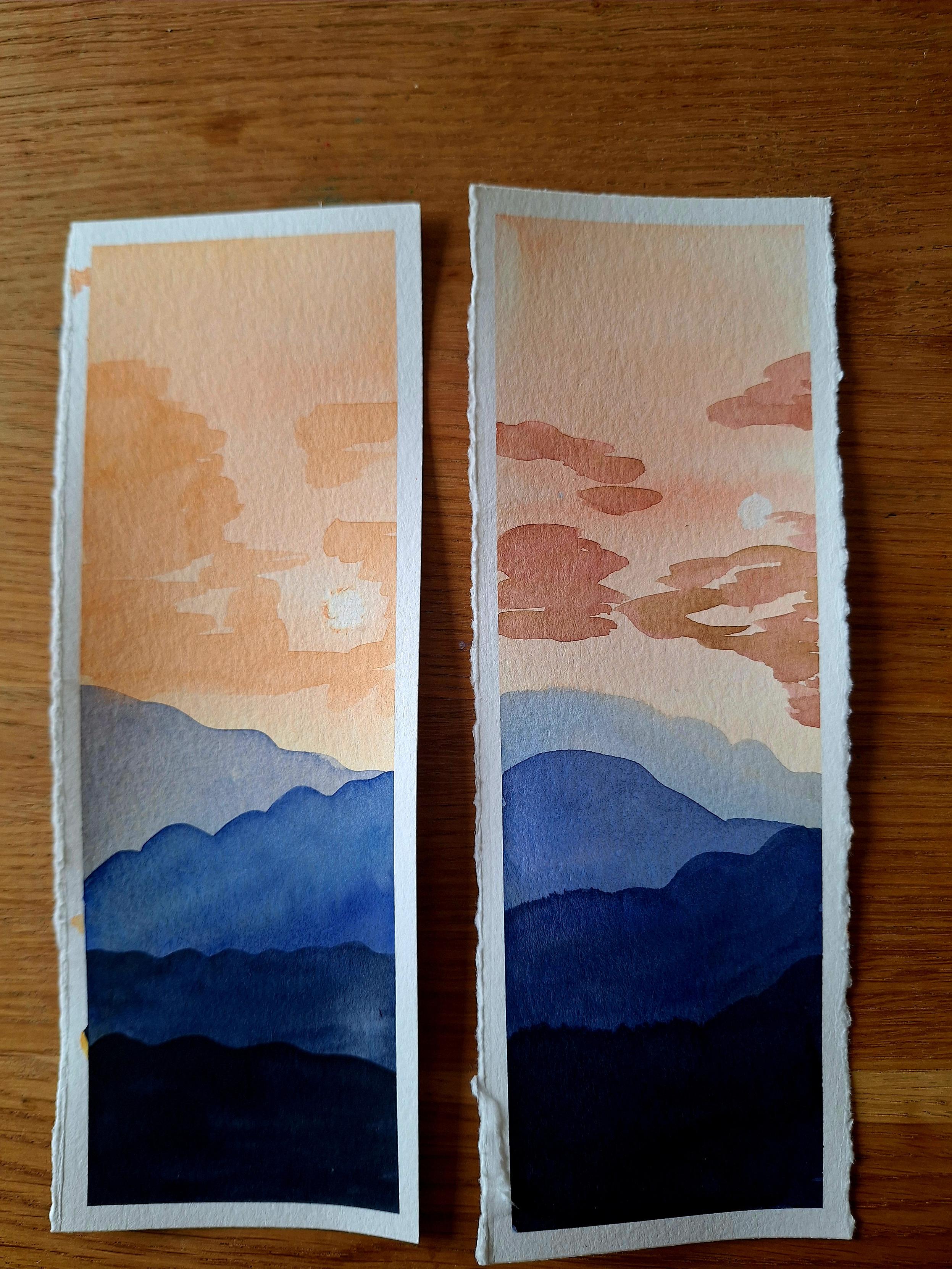 Das Bild zeigt zwei handgemalte Lesezeichen mit einer Berglandschaft im Aquarellstil auf einem Holztisch.